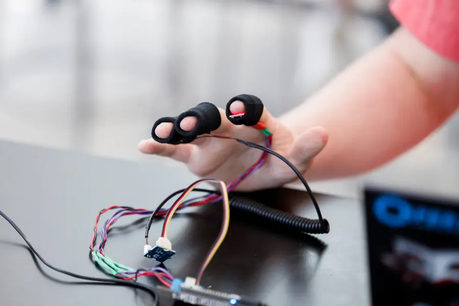 A hand rests on a table with wires connected to three fingers, the wires extend across the table and connect to a box cut off from the frame.