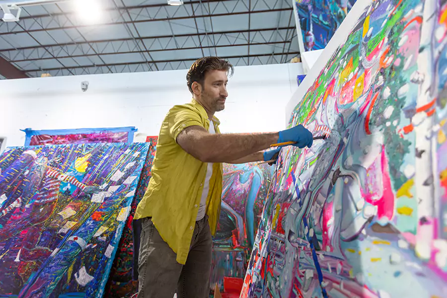 A man surrounded by paintings holds a paint brush to a canvas.