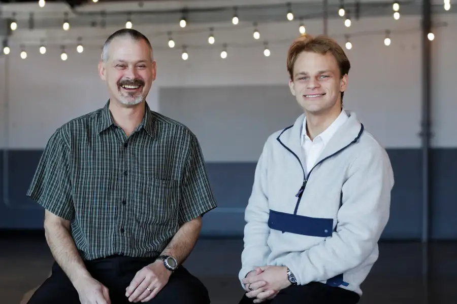 A bearded male professor in casual wear poses for a news story photo with a younger man wearing a business shirt under a quarter-zip hoodie.