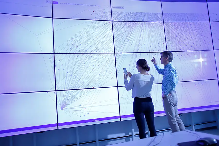 A male professor with glasses and gray hair gestures across an interactive screen wall while explaining a concept to a female student.
