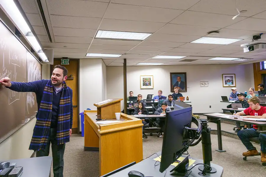 A male professor in a jaunty blue tartan scarf writes on a chalkboard while students take notes behind him.