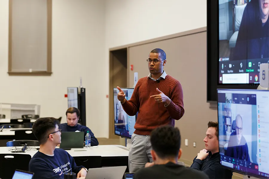 A male professor in a burnt orange sweater and glasses commands a huddle of male students looking up from laptops.