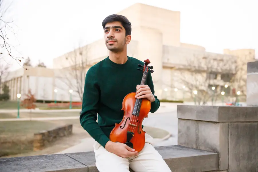 A man sits on a limestone wall while holding a violin in his lap, he looks off into the distance.