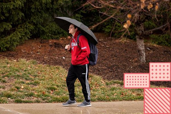 A student walks by himself in the rain. He is holding an umbrella and carrying a back pack.